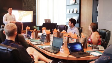 6 Menschen sitzen um einen Verhandlungstisch vor ihren Laptops und hören einer Präsentation zu